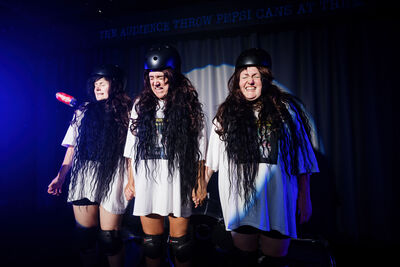 A picture of three performers on stage wearing helmets and holding hands as they brace for impact while the audience throws cans at them
