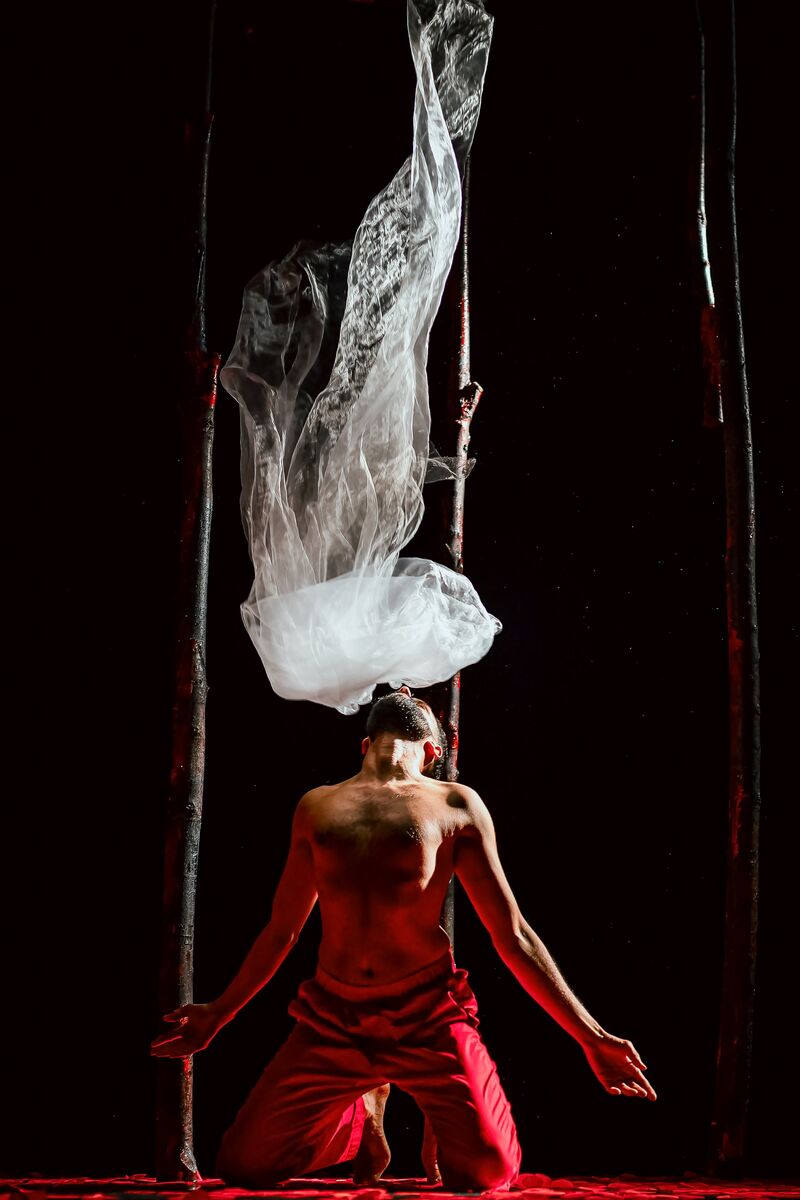 A topless man in red trousers looks toward the sky, right above his nose is some clear material, floating