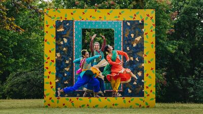 Three dancers dressed in bright Indian costumes pose inside brightly coloured, giant frames