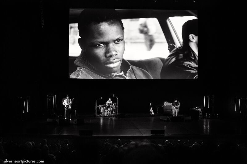 Black and white photo of a stage with 3 musicians on and a film projected onto a creen above that shows a black man staring off into the distance