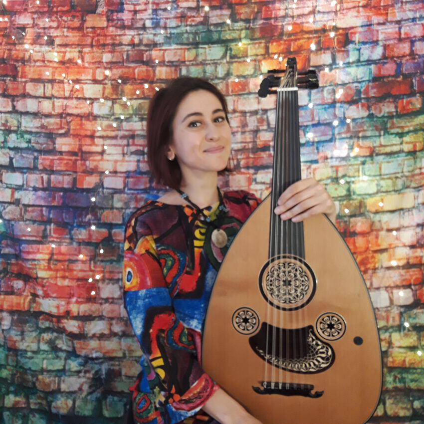 Rihab Azar holding an oud, wearing a swirly multi-coloured dress with dark red-brown hair in a bob. Background is colourful red, blue and green brick wall