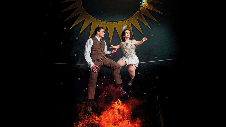 The main image is two performers sat on a raised tight wire in a dark circus tent. The circus tent is decorated with gold stars and bunting. To the right is the main character, Krista who is a young white woman of short stature with short brown hair looking away into the distance. To the left is Gerhard, a young white man with short brown hair, who gazes intently at the other performer, Krista, as he reaches his hand to touch her. Both characters’ costumes are in the style of the 1930s. Hers is a shiny silver beaded corset with white silk shorts, tights and black shoes. He looks elegant in brown trousers and a brown waistcoat over a long-sleeved white shirt and black shoes. Underneath the two performers burns a small fierce fire with red flames, reaching the soles of their feet and signifying the danger beneath this love story