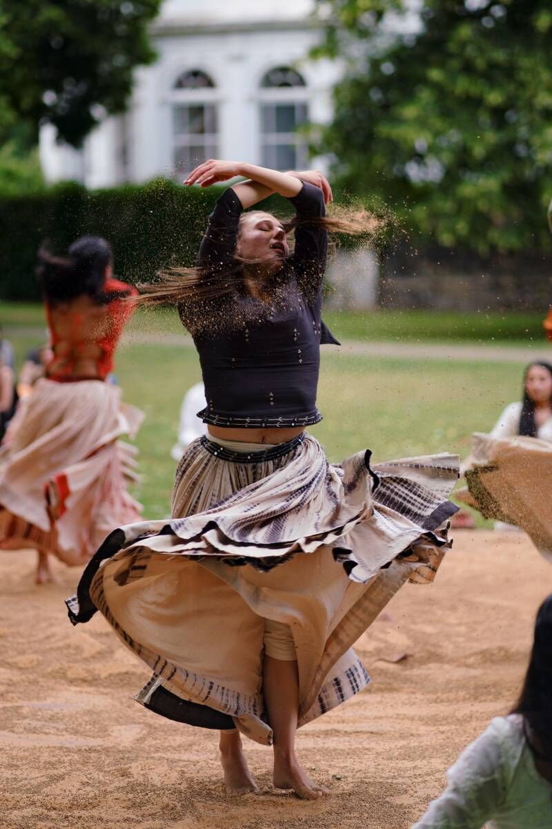A dancer spins with her arms in the air, her hair and skirt whipping around with some sand flying up around her