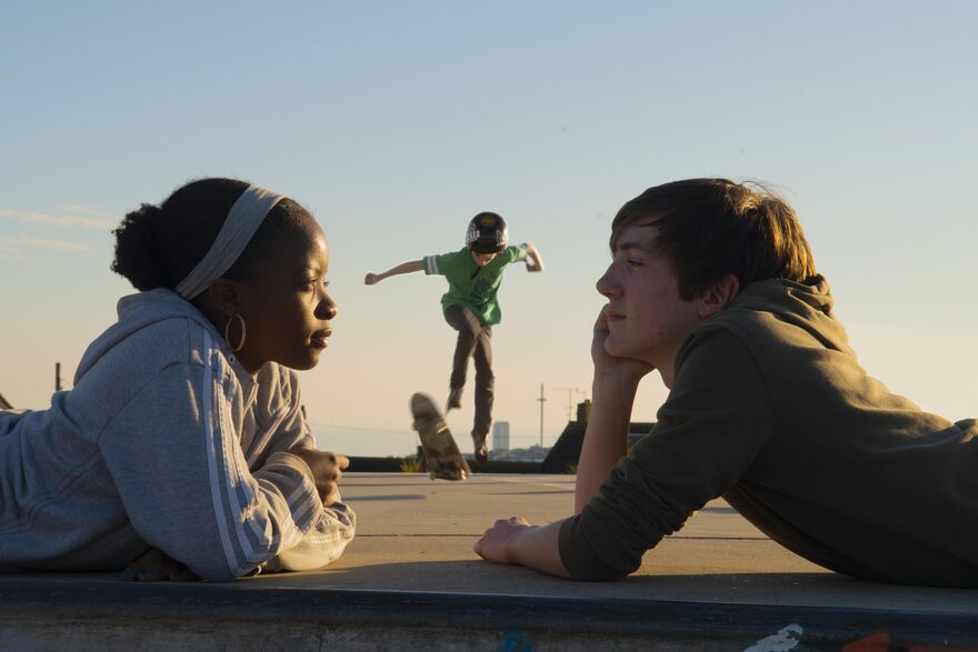 Two young people laying on their fronts gaze in to each others eyes, behind them is a skateboarder doing a trick