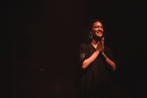 Anoushka Shankar stands smiling against a black background, her hands in a position of prayer or gratitude.