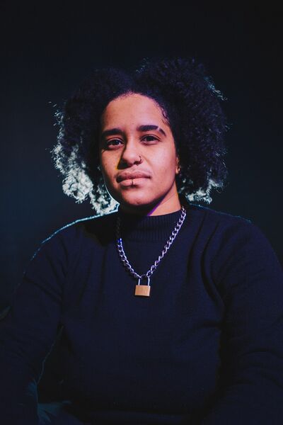 A queer person of colour looks straight ahead. They are wearing a black jumper, a silver chain with a padlock at the end, and have a nose and lip piercing. There is a light shining behind them, lighting up the ends of their hair.