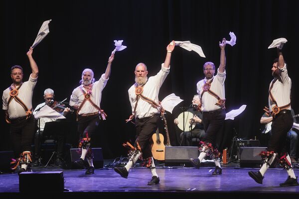 A row of Morris Men dancing on a stage, they are waving white hankies in the air