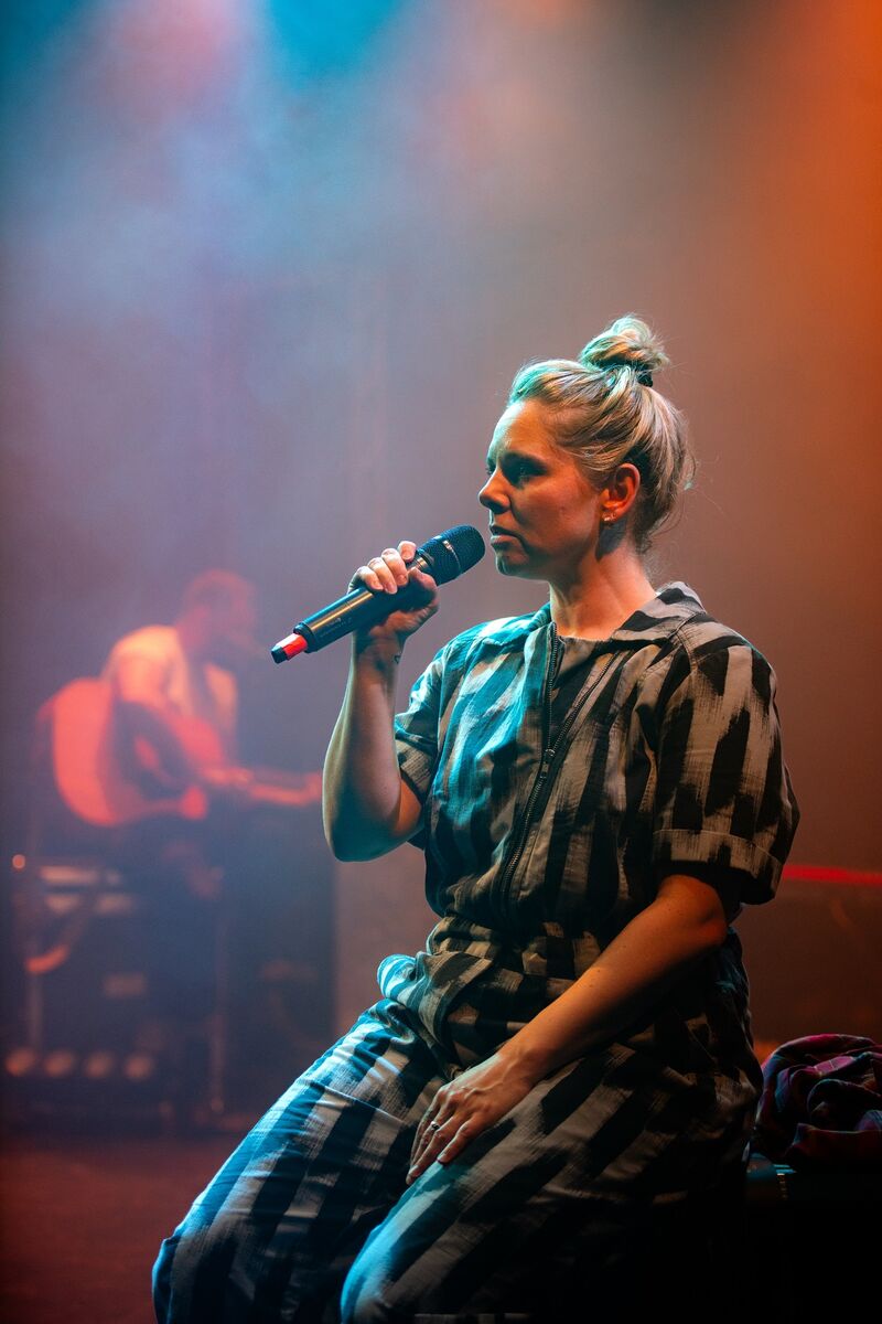 A white woman with blonde hair in a 'bun' sits whilst singing in to a wireless microphone