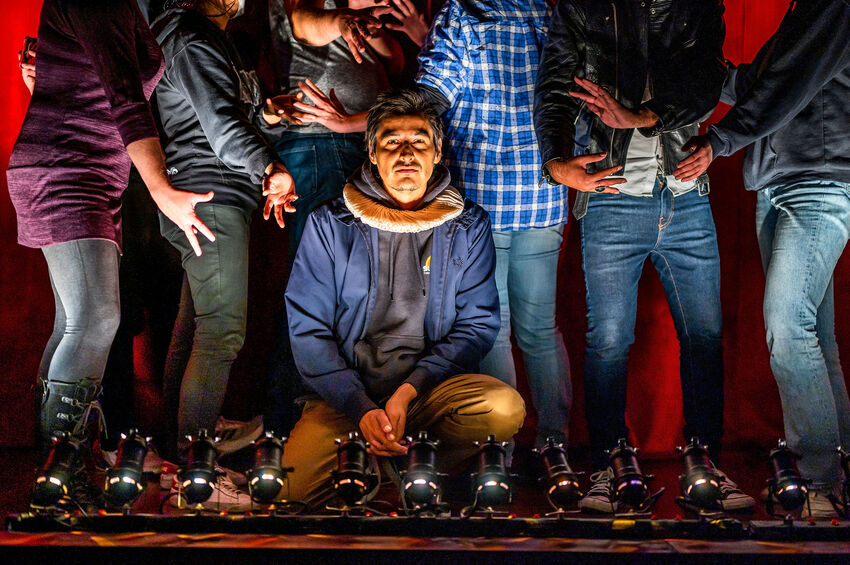 Cliff Cardinal crouches at the front of a stage wearing a hoodie and a neck ruff, people stand gesturing behind him