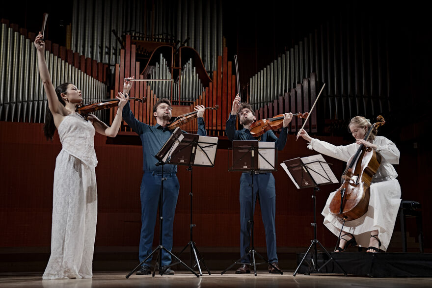 Novo Quartet on stage in front of an organ. They all have their bows in the air.