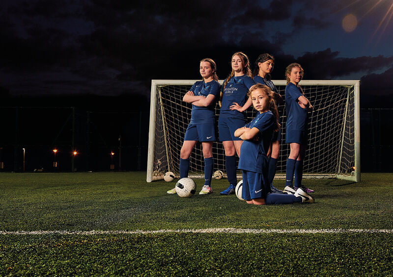 Five girls posing in front of a goal, one girl is on her knees, with her arms folded, the others are standing