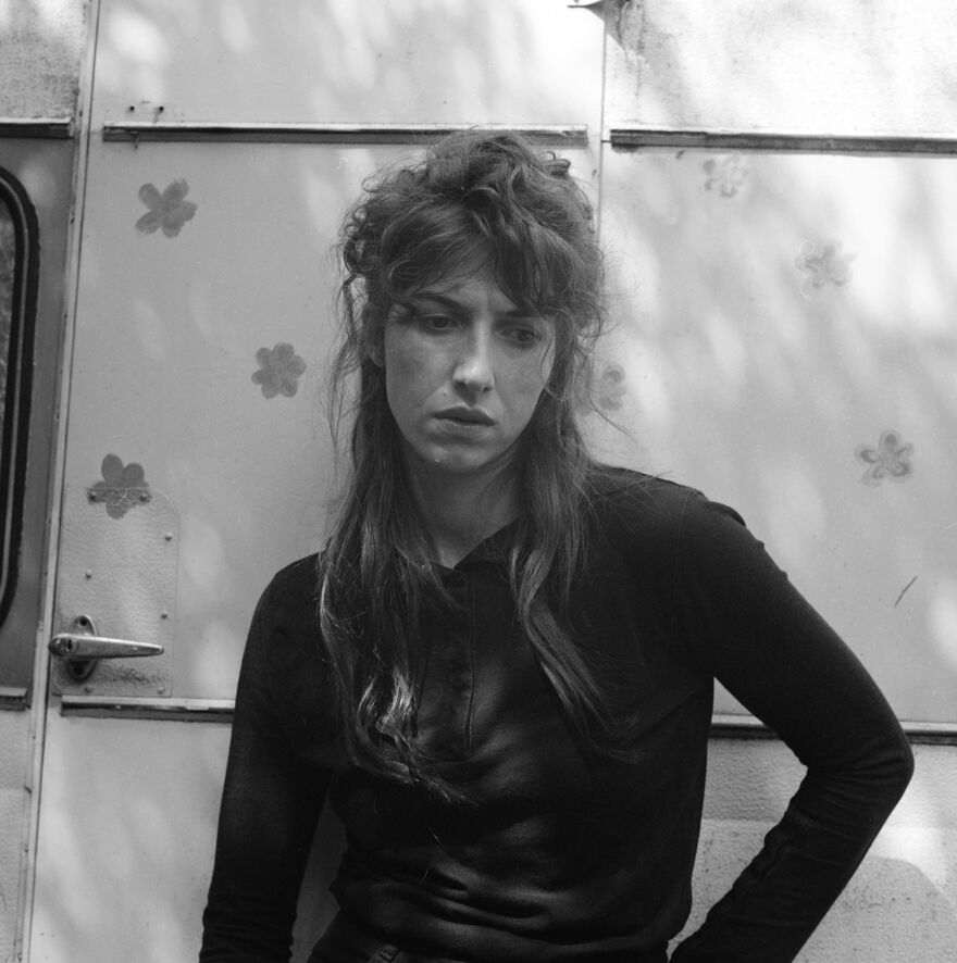 Black and white photo of Aldous Harding, a young white woman with long hair that's messily tied up in a half up half down. She's wearing a black long sleeved top and looking at the ground pensively