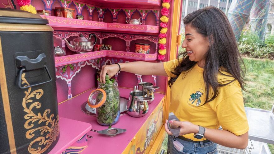 A lady in a bright yellow teeshirt pulls mint leaves out of a pot. The background is a bright pink tuk tuk adorned with tea pots and other tea making accessories.