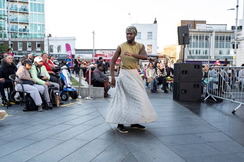 A man mid-movement wearing a cream quilted skirt and beige tanktop, audiences sat behind watching him