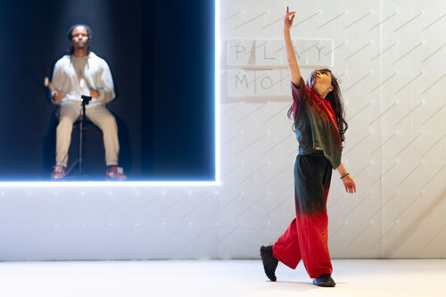 A young girl reaches her arm up to the ceiling. A man is sat in the wall behind her in a fluorescent frame playing a drum