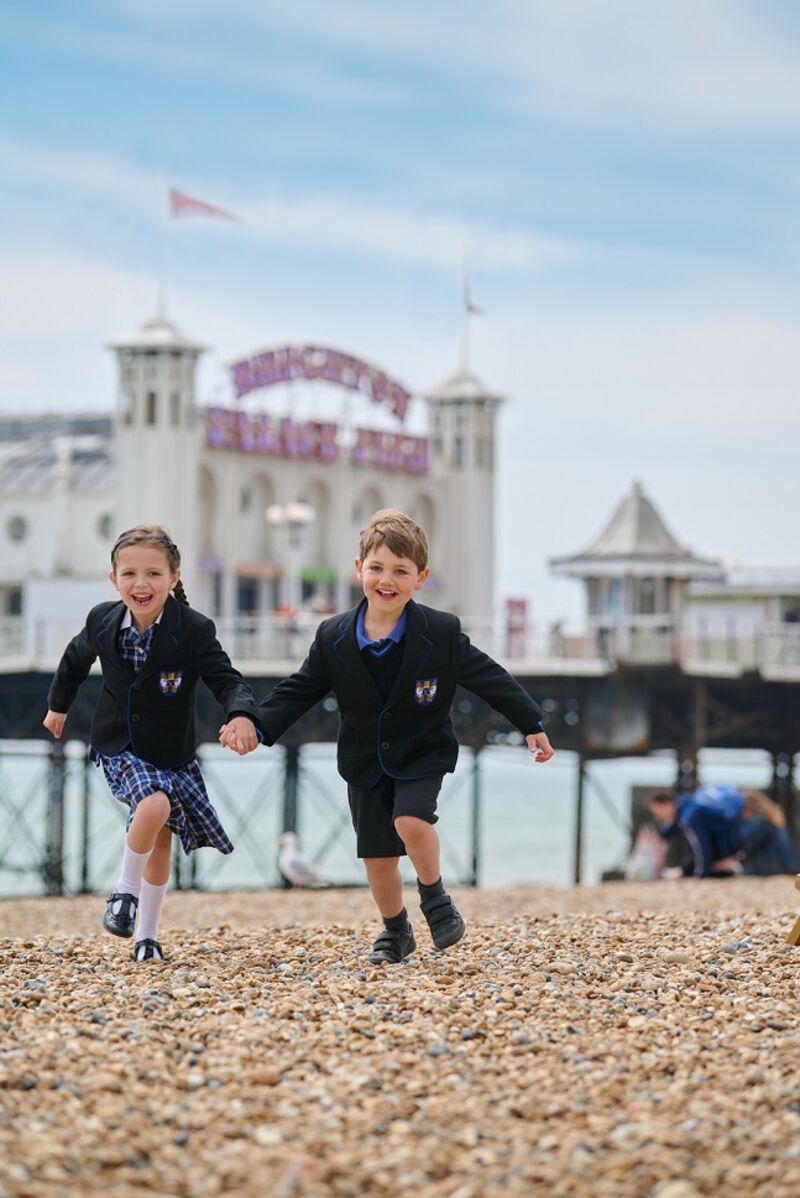 Two school children running on pebbles whilst holding hands, behind them is the Brighton Palace Pier