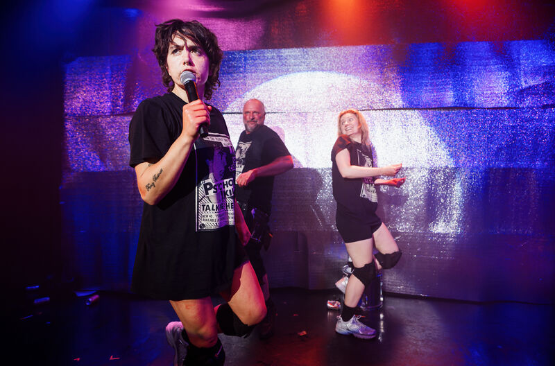 On stage picture of three performers, one is in the foreground and speaking into a microphone whilst the other two dance in the background