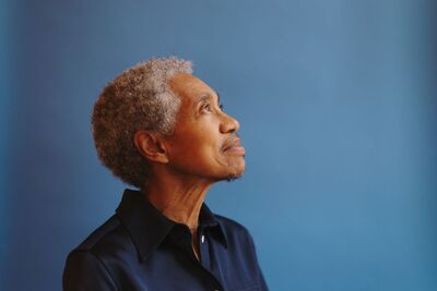 Beverly Glenn Copeland, a Black transgender man with short curly gray hair stands sideways on looking to the ceiling in front of a blue black drop