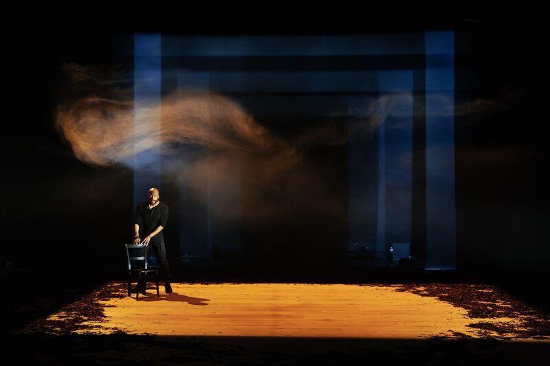 Arinze, a black man, stands behind a black chair, his hands resting on its back. Around and above him is an orange light, the air filled with smoke