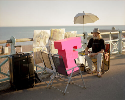 Man skerching onm seafront with a chair infront of him, there is a brighton festival logo in 3d resting on the chair