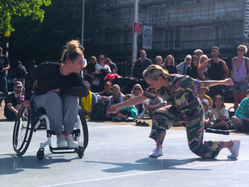 Two dancers perform together, as a crowd watches on. They are outdoors. One performer bows to the other performer, who dances in a wheelchair.