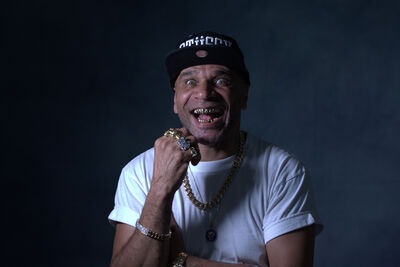 Goldie wears a white t-shirt, black hat and gold necklace and rings, smiling showing his gold teeth