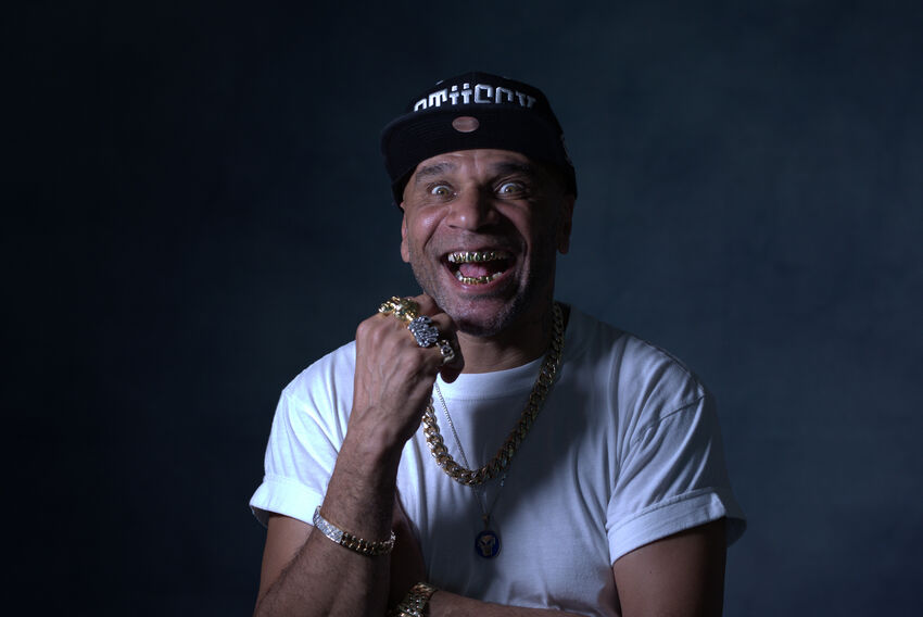 Goldie wears a white t-shirt, black hat and gold necklace and rings, smiling showing his gold teeth