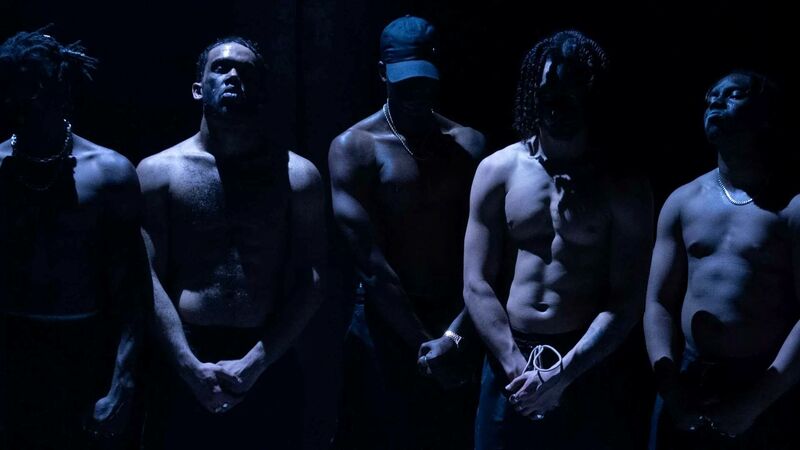Five intimidating topless men stand on a stage lit by a white light which casts shadows across their chests.