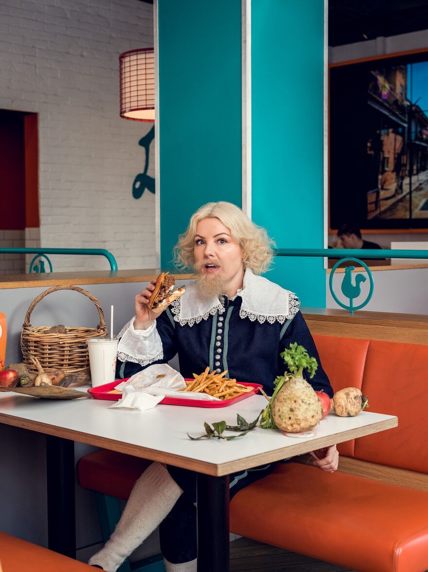 A portrait of Victoria Melody. She is sat in a modern cafe setting eating fast food, but dressed in Stewart era clothing with vegetables and wicker baskets on the table