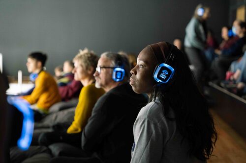 The camera points from the side of the audience, showing everyone wearing headphones that are lit up blue. The audience look towards the stage intently