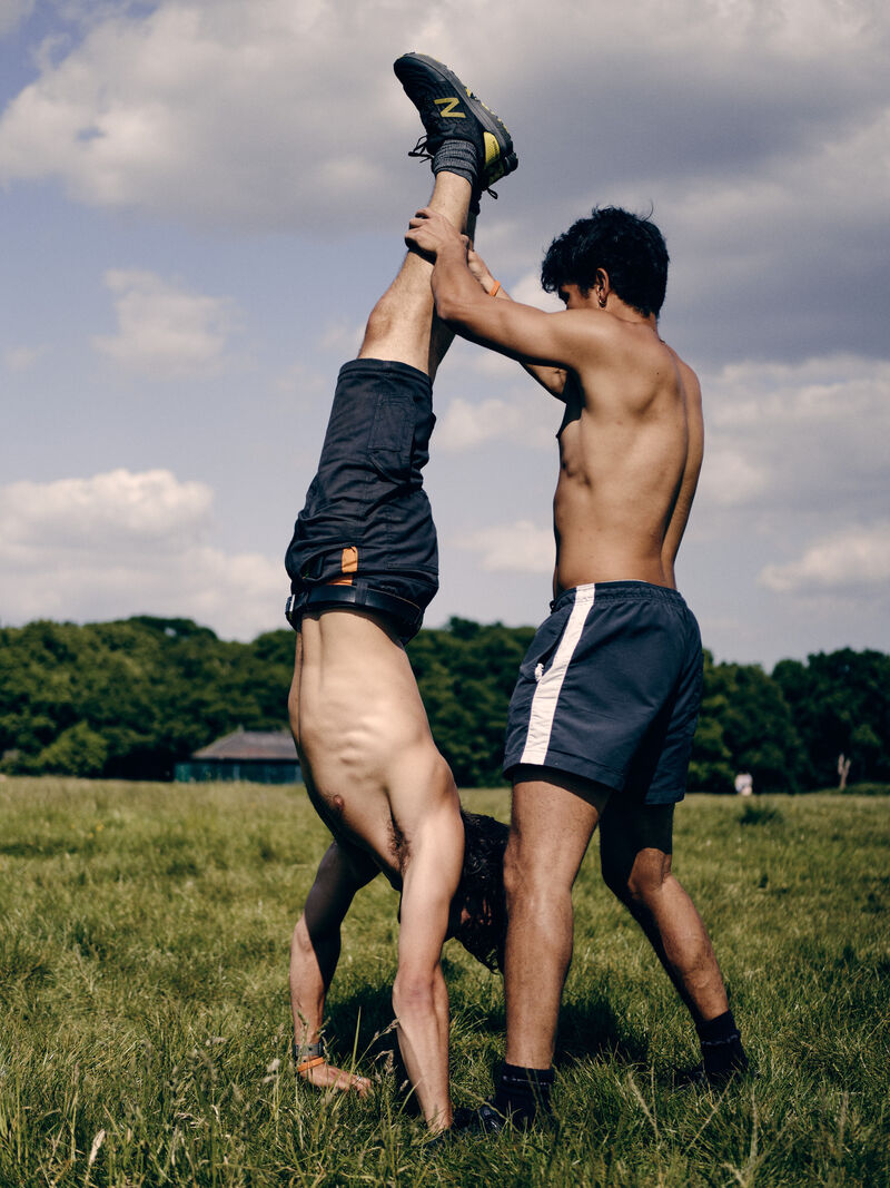 In a field of green grass, one person does a handstand, the other holds their legs. both have dark hair, blue shorts, trainers and no tops