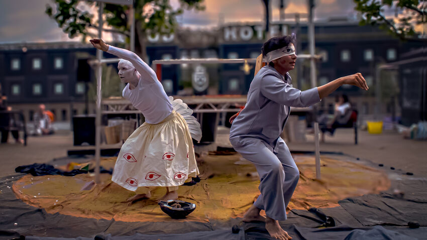 Two dancers on an outdoor, flat stage. One is all in white, the other blindfolded and in a light blue outfit. They are turned away from eachother with their left arms both held aloft, as if to represent the shape of an animal