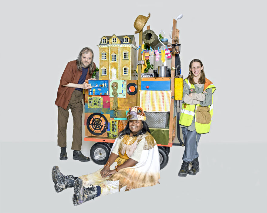 Three performers stand or sit in front of a trailer piled high with objects, including a dolls house, sun hat and yoga mat. It is very colourful