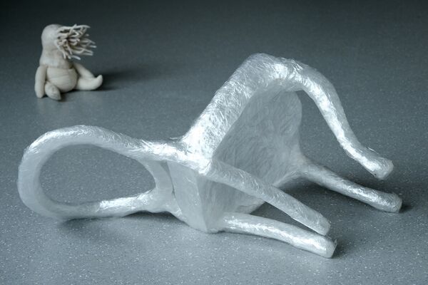 A chair made of cling film lay on its side, whilst a small alien type creature sits on the floor behind it