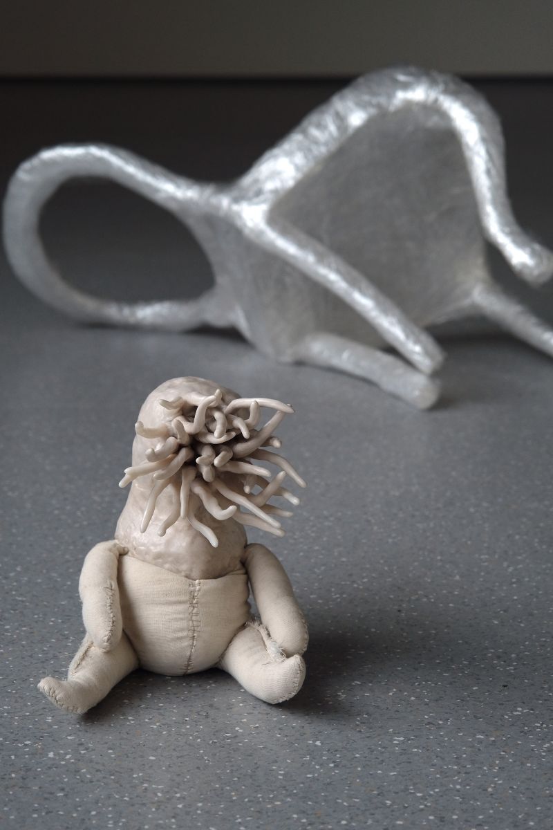 A miniature chair made of cling film lay on its side, a small clay creature sits on the floor in front of it