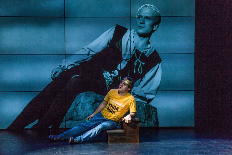 A screen showing a film with the actor lounging sideways, an actor on stage mimics this pose
