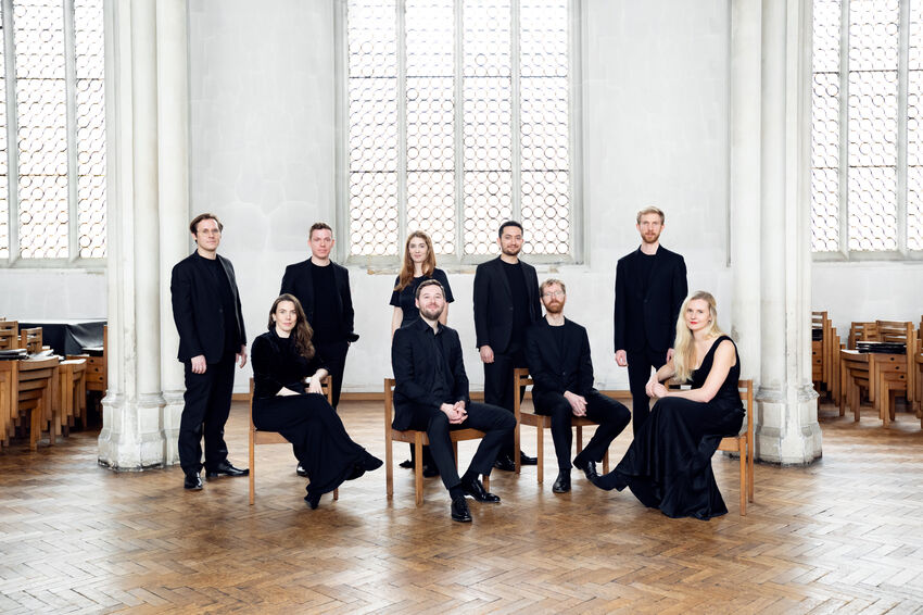 The Marian Consort, four on chairs and five standing, wear all black in a bright hall with wooden floors and white walls