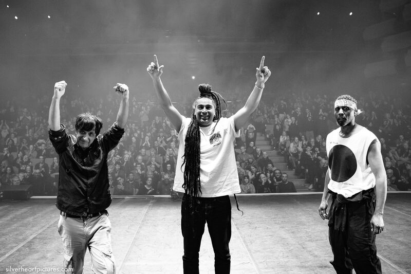 Black and white photo of three men stood on stage facing away from the audience. Left to right there's one man in a dark shirt with his arms in the air, a man with long braided hair wearing a white tshirt pointing in the air, and another man wearing a white vest and bandana 