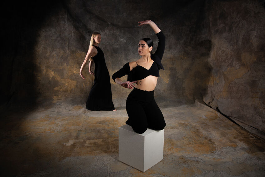 A dancer with straight blond hair and a long black dress stands in the background, in the foreground is a woman with slicked back dark hair a black crop top and trousers, kneeling on a box with her arm raised curved above her head. The backdrop is a grey/brown stone