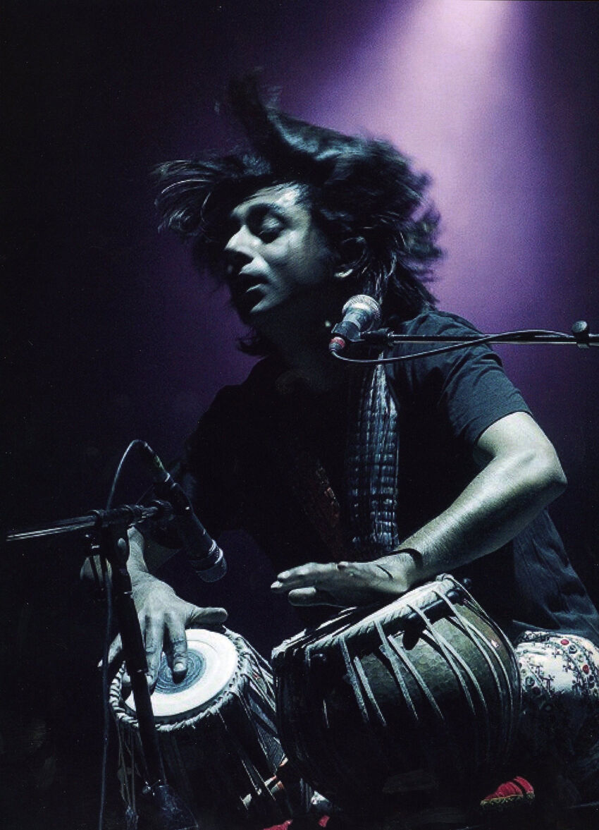 Talvin Singh playing Tabla while whipping back his hair. A purple light in the background