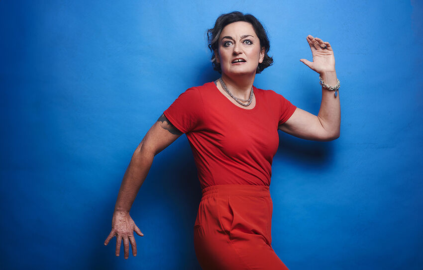 Zoe Lyons wears a red jumpsuit and stands, posing as a werewolf in front a blue wall