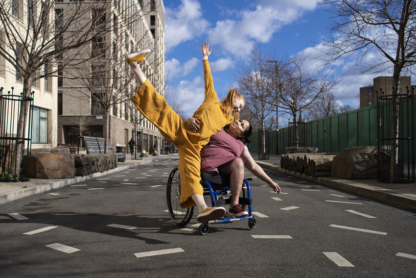 Two people dancing in the middle of the road, one is sitting in a wheelchair whilst holding the other