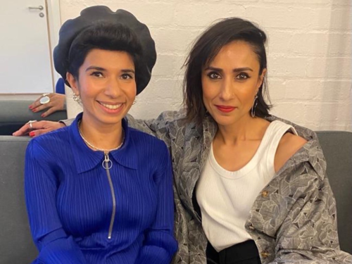 Nabihah Iqbal and Anita Rani sit next to each other on a grey sofa. Nabihah is wearing a black beret and a royal blue top and she is smiling. Anita is wearing a grey shirt and a white top. She has her arm around Nabihah.