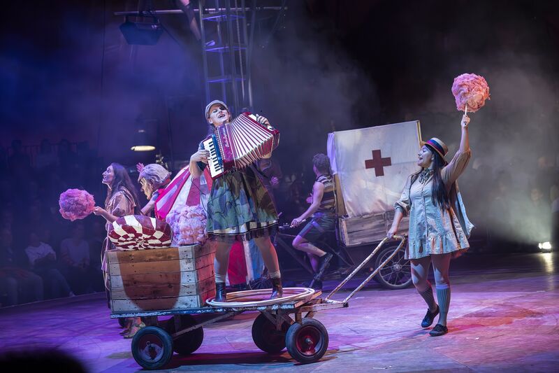 A performer in a colourful costume plays accordion while stood on a trolly with wheels. The trolly is being pushed by another performer who's holding candy floss in the air. Behind, another performer is on a bike with an ambulance tent attached