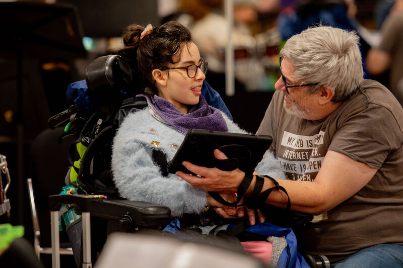 An Orchestra 360 musician sits in a wheel chair. She is making music using an iPad and is looking at the older person who is holding the iPad for her