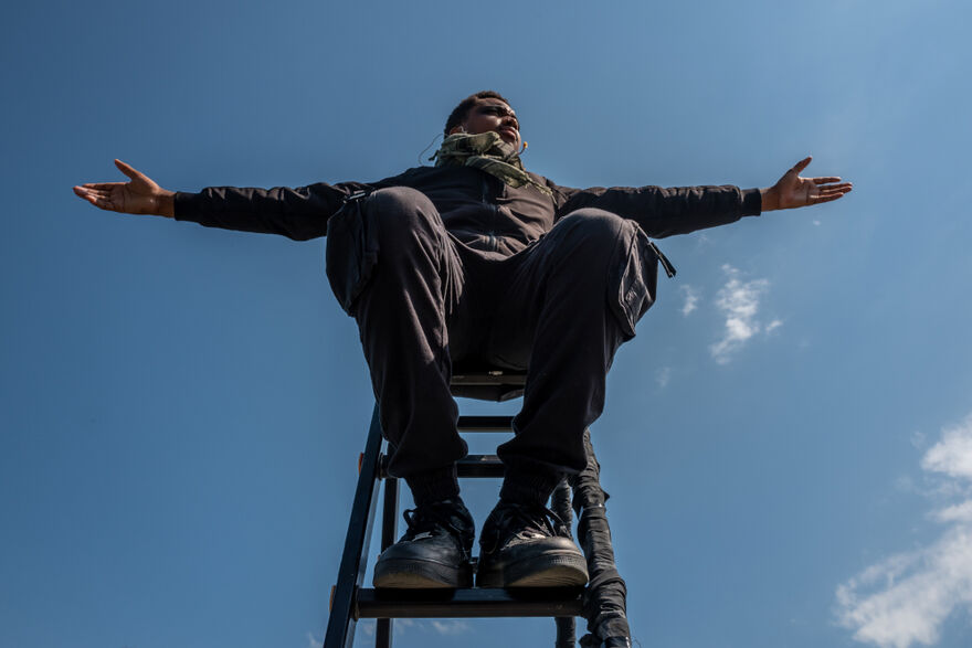 A teenage boy sits atop a ladder. He is photographed from below and has his arms outstretched against a vivid blue sky.