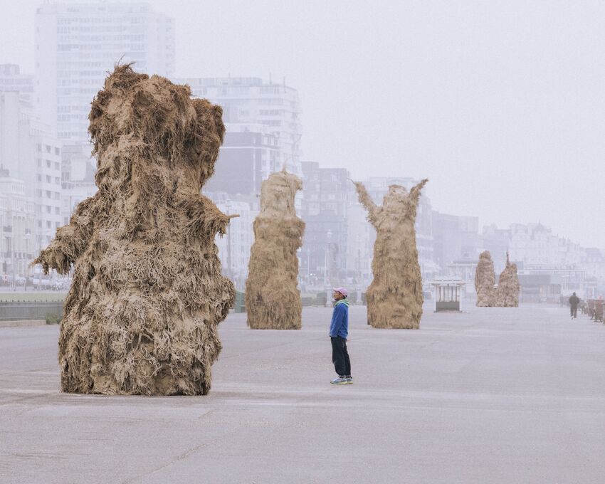 Ivan, an artist in a blue jacket, stands on Hove Promenade looking up at his Soft Machines, monumental forms made of a hay-like substance