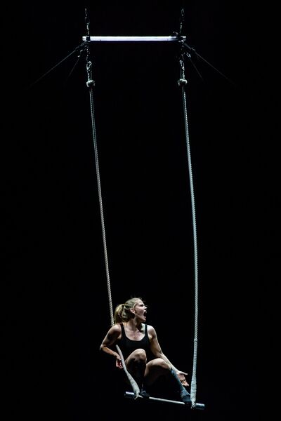 A woman in a black leotard sits on hanging bar held with aerial ropes 