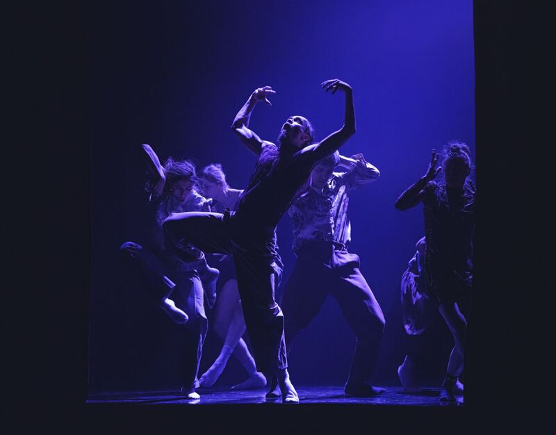  A group of 5 dancers move erratically together, each posing with a different shape and posture. The dancer at the forefront of the image is facing outwards, with his left leg planted on the ground and his right leg held high out to the side with his knee bent. His arms are curved outwards around his head, his fingers splayed, while his mouth and eyes are held wide open. The dancers are wearing an assortment of different colourful shirts, trousers and dresses.