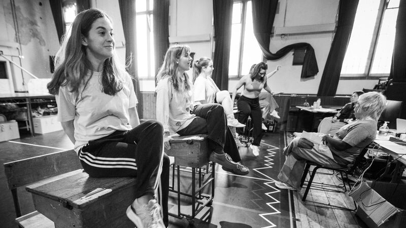 Three girls in a line sitting up straight on school desks with their legs crossed and arms behind their backs